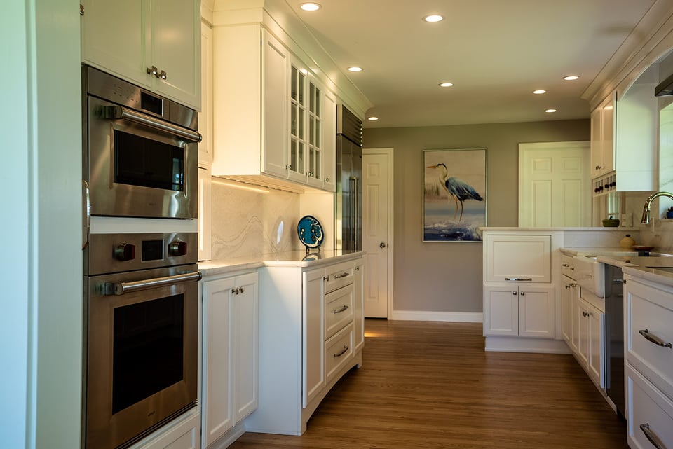Long view of a New Hampshire kitchen with white cabinets, wall oven, and soft recessed lighting above hardwood floors