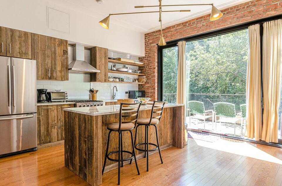 Rustic Kitchen With Wood Flooring and Cabinetry. Countertop Extends With Two Chairs Across From Stainless Steel Refrigerator and Oven